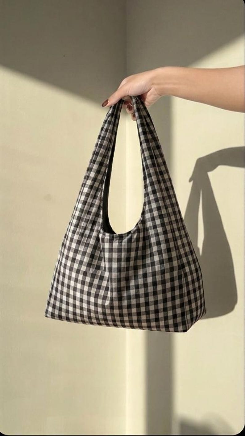 May include: A hobo-style shoulder bag featuring a classic black and white gingham check pattern. The bag has a wide, integrated shoulder strap and a spacious, unstructured body with a flat base, constructed from a woven fabric material.
