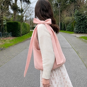 May include: A soft pink shoulder bag featuring a large, tied bow detail on the strap. The bag is made of a textured, ribbed fabric and is worn over a cream-colored long-sleeved sweater paired with a white skirt featuring a small floral print.