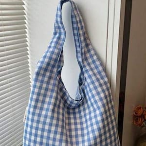 May include: A soft hobo-style shoulder bag featuring a light blue and white gingham checkered pattern. The bag has a single integrated shoulder strap and a slouchy, unstructured silhouette that hangs vertically with a wide opening at the top.