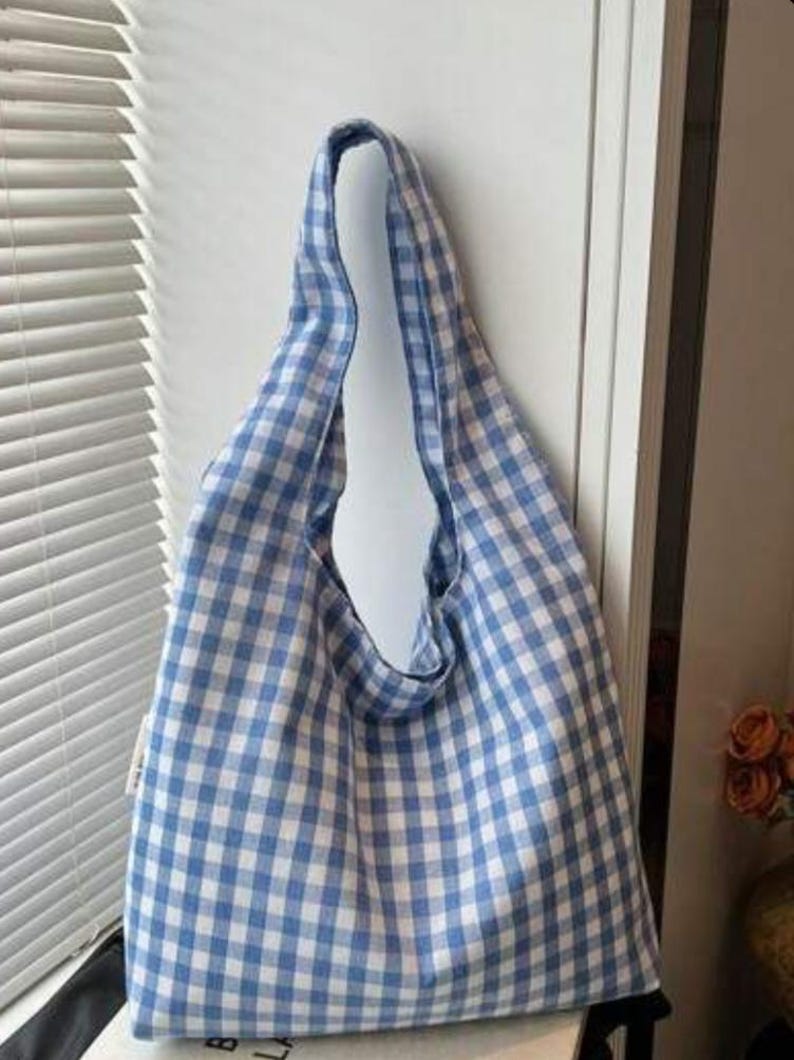 May include: A soft hobo-style shoulder bag featuring a light blue and white gingham checkered pattern. The bag has a single integrated shoulder strap and a slouchy, unstructured silhouette that hangs vertically with a wide opening at the top.