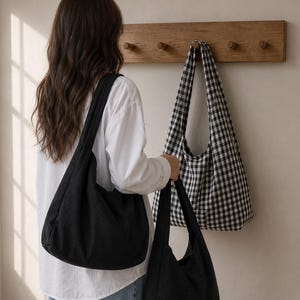 May include: A solid black hobo bag with a wide shoulder strap and a matching black hobo bag held in the hand, alongside a black and white gingham checkered hobo bag hanging on a wooden wall hook rack, all featuring a soft, unstructured fabric design.