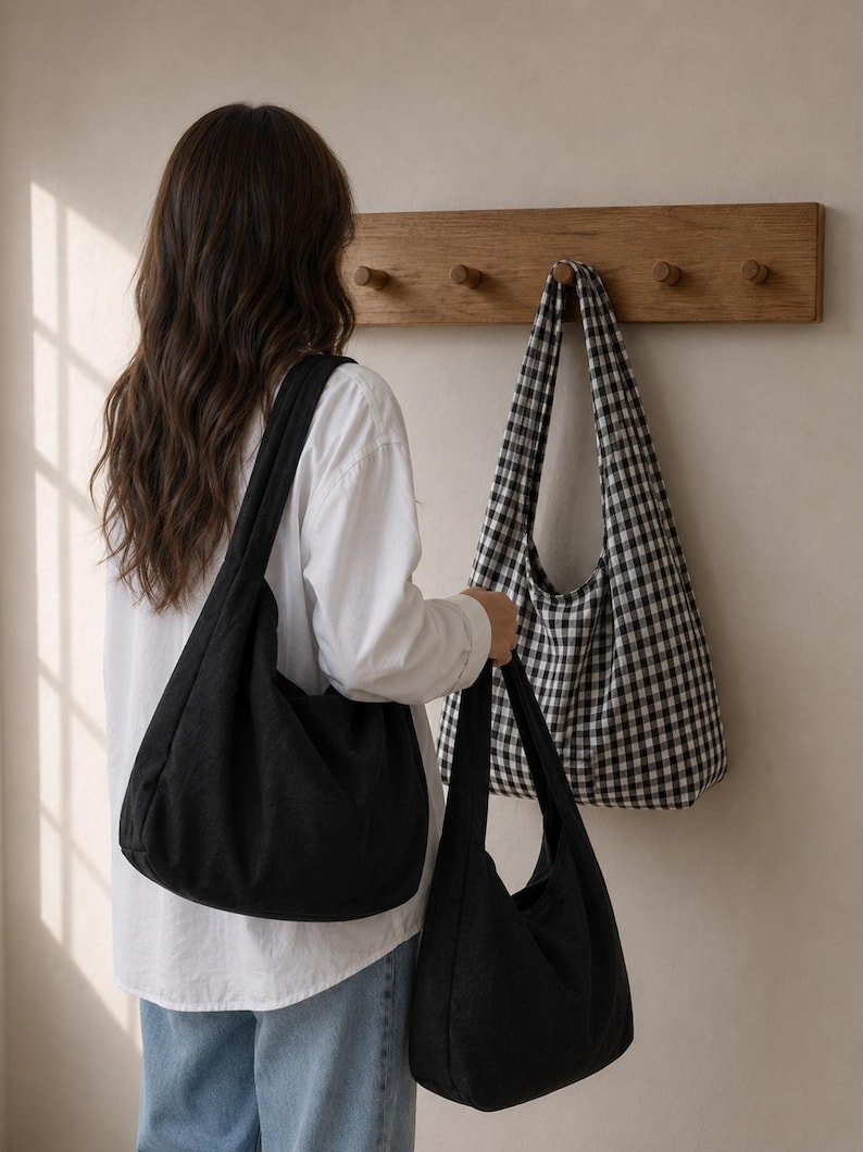 May include: A solid black hobo bag with a wide shoulder strap and a matching black hobo bag held in the hand, alongside a black and white gingham checkered hobo bag hanging on a wooden wall hook rack, all featuring a soft, unstructured fabric design.