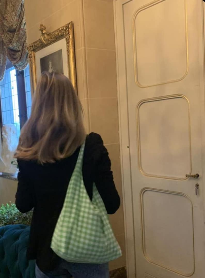 May include: A light green and white gingham patterned hobo bag with a soft, unstructured shape and a wide shoulder strap, worn over a black long-sleeved top and blue denim jeans, showcasing a casual and functional accessory design.
