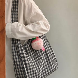 May include: A black and white gingham patterned fabric tote bag with a wide shoulder strap. A small, round, plush pink character charm with a green leaf accent is attached to the side of the bag, featuring a simple embroidered face.