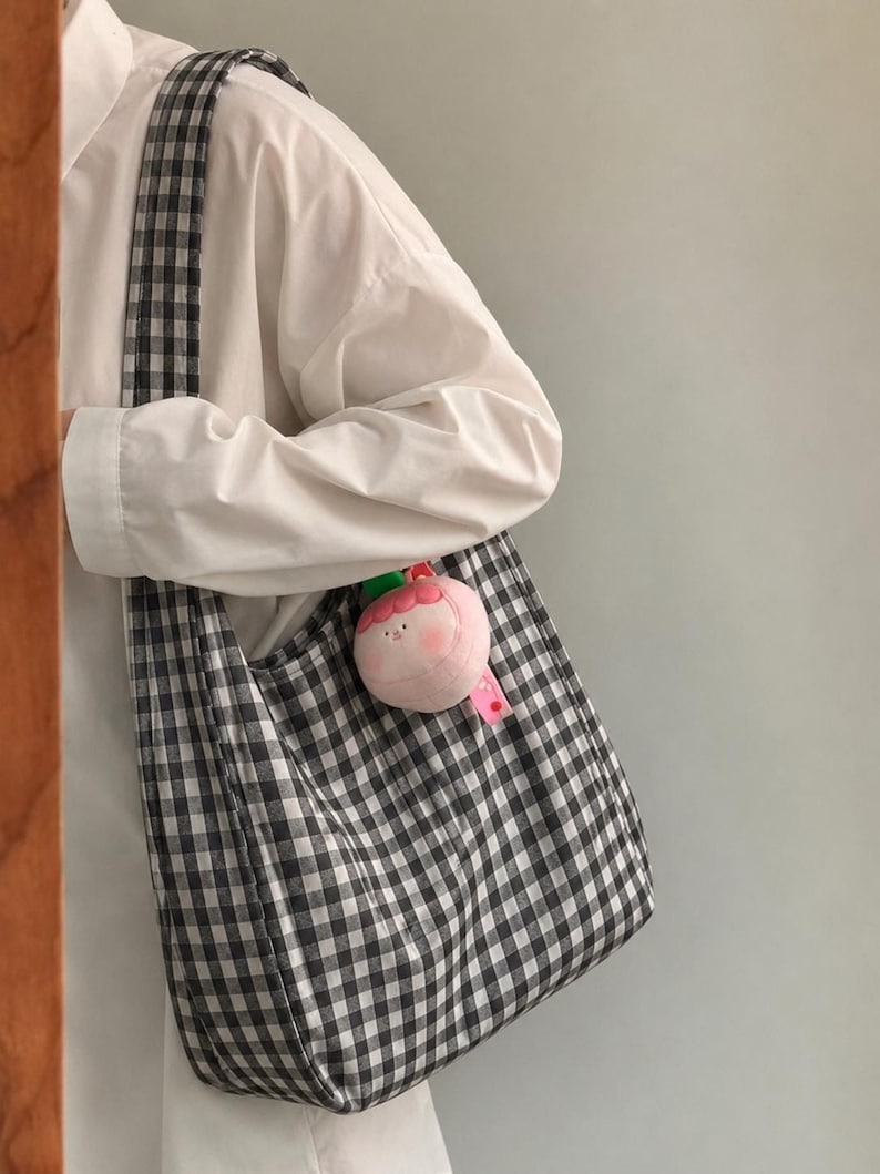 May include: A black and white gingham patterned fabric tote bag with a wide shoulder strap. A small, round, plush pink character charm with a green leaf accent is attached to the side of the bag, featuring a simple embroidered face.