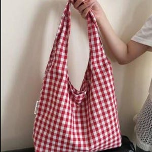 May include: A soft fabric hobo bag featuring a classic red and white gingham checkered pattern. The bag has a wide, integrated shoulder strap and a rectangular body shape. A small white fabric tag is visible on the side seam of the bag.