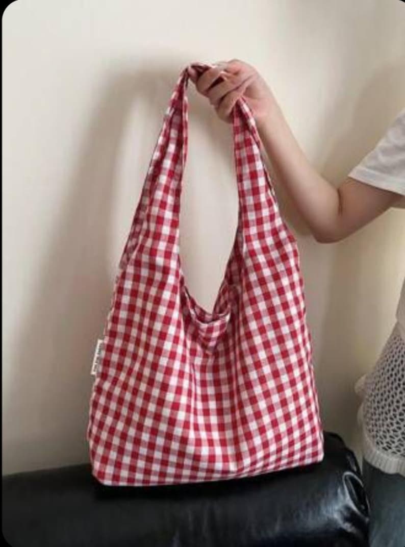 May include: A soft fabric hobo bag featuring a classic red and white gingham checkered pattern. The bag has a wide, integrated shoulder strap and a rectangular body shape. A small white fabric tag is visible on the side seam of the bag.