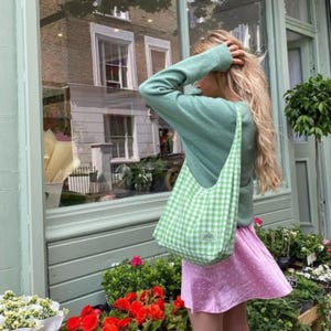 May include: A light green long-sleeved sweater paired with a light green and white gingham shoulder bag featuring a small rectangular label. The outfit includes a light pink skirt with a subtle white pattern, worn by a person with long blonde hair.