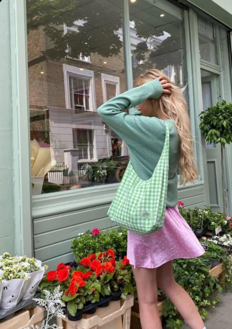 May include: A light green long-sleeved sweater paired with a light green and white gingham shoulder bag featuring a small rectangular label. The outfit includes a light pink skirt with a subtle white pattern, worn by a person with long blonde hair.
