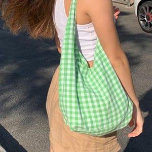 May include: A soft, unstructured hobo-style shoulder bag made from fabric featuring a bright green and white gingham checkered pattern. The bag has a wide, integrated shoulder strap and a spacious, rounded body that hangs at the side.