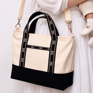 May include: A cream-colored canvas tote bag with a solid black base and two black handles. The bag features vertical black ribbon straps decorated with a repeating white bow pattern, a detachable shoulder strap, and a horizontal front pocket.
