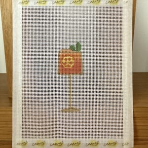 May include: A needlepoint canvas featuring a square orange cocktail glass design with a yellow citrus slice and green mint leaves. The design is centered on a white grid mesh with taped edges that have the word canvas printed repeatedly in yellow.