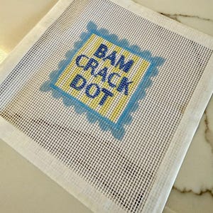 May include: A square needlepoint canvas featuring a yellow center square with a blue scalloped border. The text BAM CRACK DOT is printed in blue block letters in the center. The canvas has a white grid pattern throughout the entire square piece.