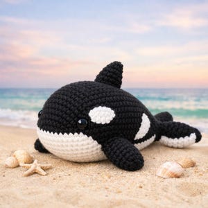 Peut inclure: Une peluche d'orque faite à la main au crochet. L'orque est noire et blanche, avec un ventre et des taches blanches. Elle est posée sur une plage de sable avec des coquillages et une étoile de mer. L'arrière-plan montre l'océan et un ciel coloré.