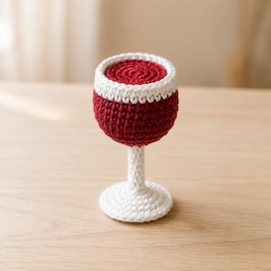 May include: A handcrafted crochet wine glass featuring a deep red bowl with a white rim and a white stem and base. The texture is created with visible yarn stitches, forming a rounded cup shape that transitions into a slender stem and circular foot.