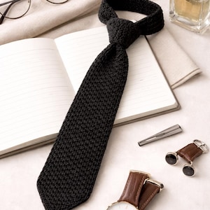 May include: A black knitted tie is knotted and lies on an open notebook. A gold-toned watch with a brown leather band, eyeglasses, a bottle of cologne, a tie clip, and cufflinks are also present. The items are arranged on a light-colored surface.