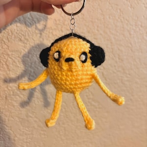 May include: Yellow crocheted keychain featuring a cartoon character with black ears and eyes. The keychain is round with small arms and legs. The character is attached to a silver keyring.
