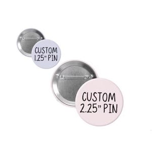 May include: A collection of custom pin-back buttons in various sizes. The pins are round with a metal backing and a white or light pink face. The text "CUSTOM 1.25" PIN" and "CUSTOM 2.25" PIN" is printed on the faces.