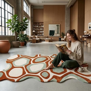May include: An irregular, organic-shaped area rug featuring a bold, abstract pattern of interlocking shapes in cream, terracotta orange, and sage green. The rug has a thick, tufted texture with defined borders between the contrasting color blocks.
