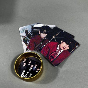 May include: A collection of BTS merchandise, including photo cards and a small, gold-colored container. The photo cards feature images of the band members in various poses. The container has a black and white image of the band with the letters "BTS" on it.
