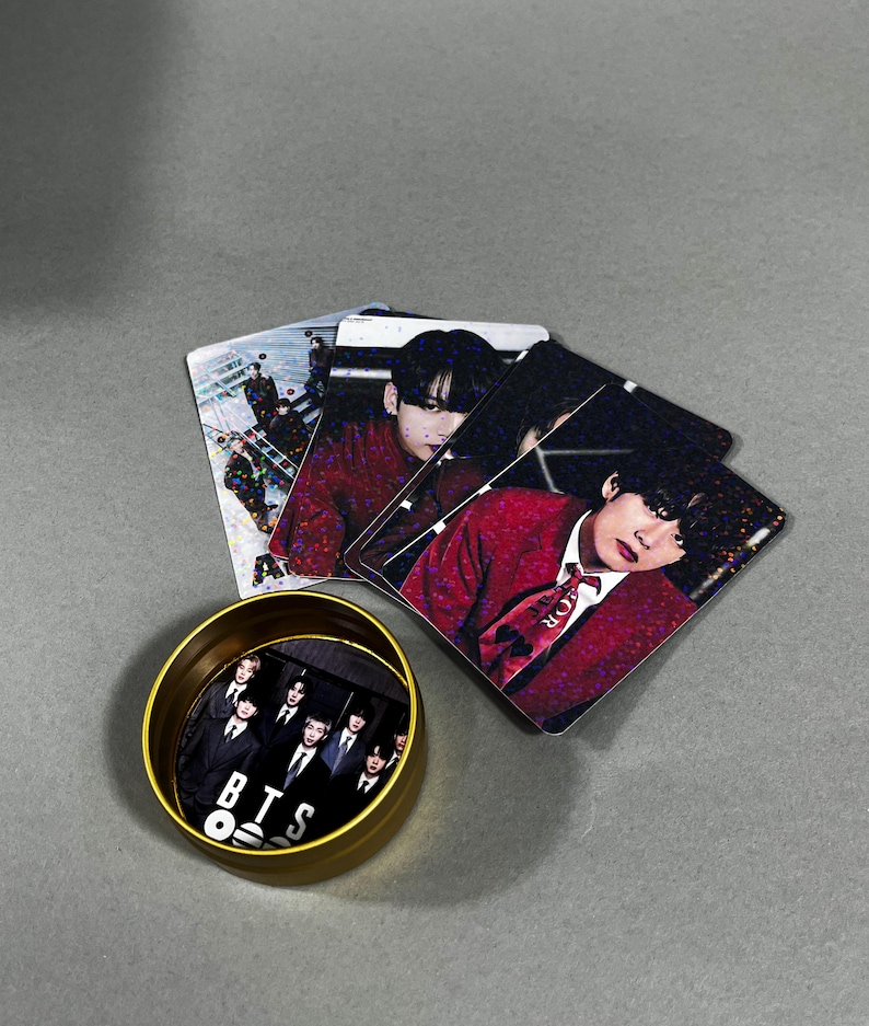 May include: A collection of BTS merchandise, including photo cards and a small, gold-colored container. The photo cards feature images of the band members in various poses. The container has a black and white image of the band with the letters "BTS" on it.