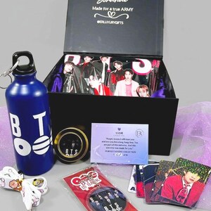 May include: A gift box with various BTS-themed merchandise. Includes a blue water bottle with white lettering, a keychain, a pin, photo cards, and a box with images of the band members. The box has the text "Borahae" and "Made for a true ARMY".