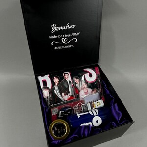 May include: A black gift box lined with purple satin containing BTS merchandise. The lid interior features the text Borahae, Made for a true ARMY, and @ERLUXURYGIFTS. Inside are photo cards, a blue tumbler, and a small circular gold-rimmed item.