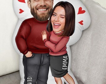 May include: A custom-designed pillow featuring a cartoon illustration of a couple. The man is wearing a maroon sweater and gray pants, with the name "DAVID" printed on his leg. The woman, wearing a matching sweater and black skirt with "EMMA" printed on it, is winking. Red hearts are scattered around.