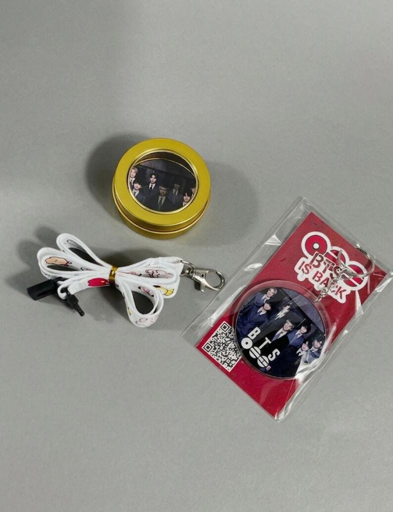 May include: A collection of BTS merchandise, including a round tin, a lanyard, and a keychain. The tin is gold with a clear window showing a photo. The lanyard is white with a printed design. The keychain is round with a photo and the text "BTS IS BACK".