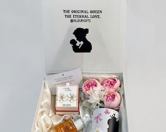 May include: A white gift box containing a Camila Coelho earring box, a bottle of amber body oil, a white mug with a black silhouette and flower print, and three pink peony flowers. The lid features the text THE ORIGINAL QUEEN THE ETERNAL LOVE.