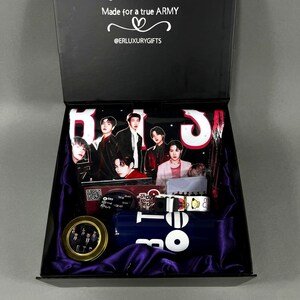 May include: A black gift box with a variety of items, including a tumbler, bracelets, and a pin, all featuring images of a musical group. The box is lined with purple fabric and has text that reads "Made for a true ARMY".
