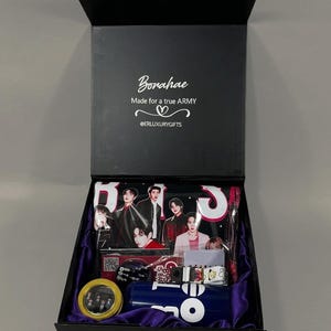 May include: A black gift box with a purple satin lining containing BTS merchandise. The lid interior features the text "Borahae Made for a true ARMY @ERLUXURYGIFTS". Items include a photo-printed pouch, a round tin, and a blue water bottle.