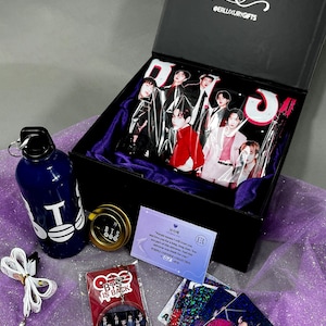 May include: A black gift box containing a BTS-themed collection, including a blue water bottle with white text, a round metal tin, a white lanyard, a circular keychain with a group photo, a purple information card, and several holographic photo cards.