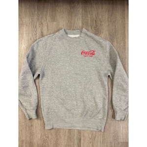 May include: A grey sweatshirt with a crew neck and long sleeves. The Coca-Cola logo and "TASTE TOUR" are printed in red on the chest. The sweatshirt is made of a soft, comfortable material.