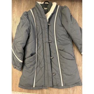 May include: A gray quilted coat with white trim and three dark buttons. The coat has a high collar and two front pockets. The coat is a mid-length style, suitable for cooler weather.