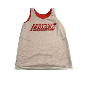 May include: A white sleeveless mesh jersey with a red interior lining. The front features red text that reads NANCY REHM INDIANA ALL STARS with star graphics integrated into the lettering. The garment has a scoop neckline and a straight hem.