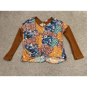 May include: A long-sleeve top featuring a V-neckline and a front twist detail. The bodice displays a patchwork pattern of blue, orange, and white floral and abstract prints, while the long sleeves are made of a solid, ribbed, tobacco-brown fabric.
