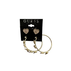 Op de afbeelding: Een sieradenset van Guess Los Angeles met twee hartvormige oorknopjes met een crèmekleurig midden en de letter G, gecombineerd met twee goudkleurige oorringen versierd met kleine vlinderbedels aan de buitenrand van de ringen.