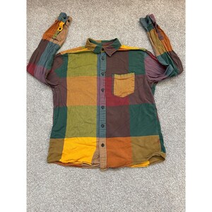 May include: A long-sleeved button-down shirt featuring a large, multi-coloured block pattern in shades of mustard yellow, forest green, deep red, and brown. The shirt includes a standard collar, a single chest pocket, and dark buttons down the front.