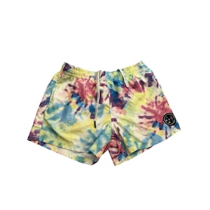 May include: Tie-dye swim shorts in yellow, pink, blue, and purple. The shorts have a drawstring waist and a small black circular patch with white text. A colourful summer garment.