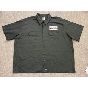 May include: A charcoal gray short-sleeve button-down work shirt featuring a pointed collar, two chest pockets with button flaps, and a rectangular patch on the left chest that reads Distribution NOW in black and red text on a white background.