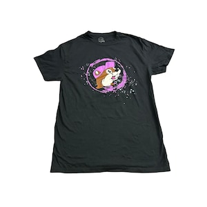 May include: A black short-sleeve t-shirt featuring a graphic of a brown cartoon beaver wearing a pink baseball cap. The beaver is centered within a circular pink brushstroke design with white paint splatters surrounding the central image.