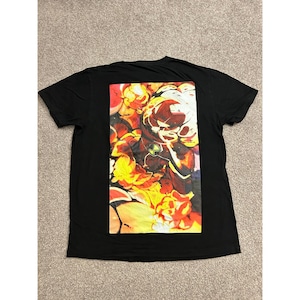 May include: A black short-sleeved t-shirt featuring a large rectangular graphic print on the back. The print displays a stylized character in a dark suit with a green circular emblem on the chest, surrounded by vibrant orange, yellow, and red flames.