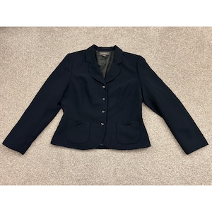 May include: A dark navy blue blazer featuring a rounded collar, a front button closure with four small buttons, and two patch pockets at the waist, each adorned with a small decorative bow detail. The jacket has long sleeves and a structured fit.