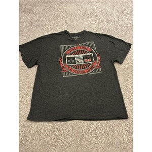 Pode incluir: Uma t-shirt cinzento-carvão de manga curta com uma impressão gráfica de um comando de videojogos retro dentro de um emblema circular vermelho e branco. O texto diz "SINCE 1985" e "OLD SCHOOL GAMER" numa fonte branca e vermelha.