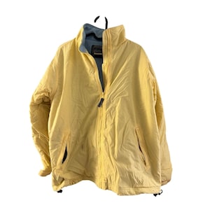 Könnte beinhalten: Hellgelbe Jacke mit durchgehendem Reißverschluss und zwei Seitentaschen. Die Jacke hat einen hohen Kragen und ein hellblaues Innenfutter. Sie hängt an einem schwarzen Kleiderbügel.