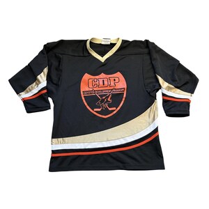 May include: Black hockey jersey with gold, white, and orange accents. Features a V-neck collar and the text "CDP Coyotes Development Program" with a coyote graphic on the front. The jersey has gold stripes on the sleeves.