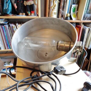 Könnte beinhalten: Eine silberne Metalllampe mit einem schwarzen Kabel und einer klaren Glühbirne. Die Lampe steht auf einer weißen Oberfläche.