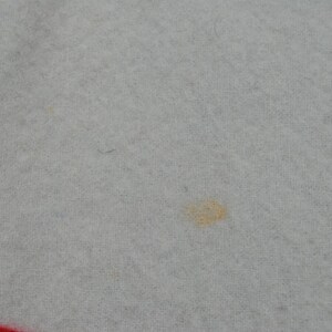 May include: A light gray fabric with a soft, fuzzy texture. There is a small, faint orange-yellow stain visible near the center of the frame. A portion of a bright red border or trim is visible along the bottom edge of the textile material.