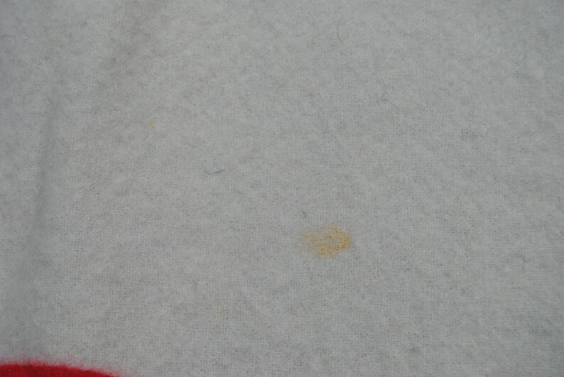 May include: A light gray fabric with a soft, fuzzy texture. There is a small, faint orange-yellow stain visible near the center of the frame. A portion of a bright red border or trim is visible along the bottom edge of the textile material.